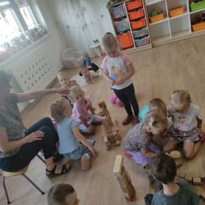 Dzieci siedzą na podłodze wraz z nauczycielem i budują z drewnianych klocków tort