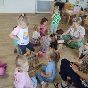 Dzieci siedzą na podłodze wraz z nauczycielem i budują z drewnianych klocków tort