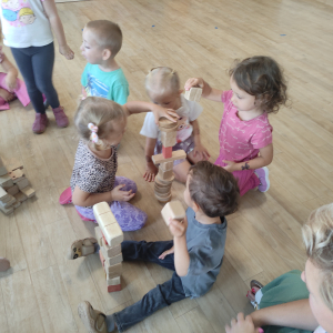 Dzieci siedzą na podłodze wraz z nauczycielem i budują z drewnianych klocków tort