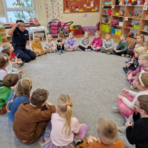 Dzieci siedzą w kole i z uwagą wysłuchują wiadomości o zasadach ruchu drogowego dla pieszych