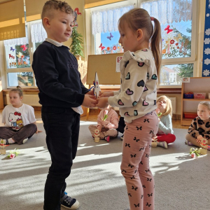 Dziewczynka składa chłopcu życzenia z okazji Dnia Mężczyzny i wręcza upominek - drewnianą rakietę z lizakiem