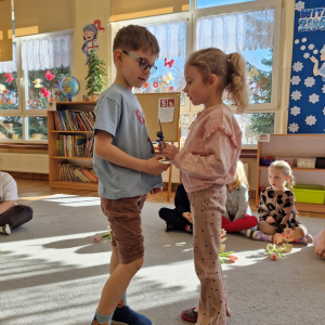 Dziewczynka składa chłopcu życzenia z okazji Dnia Mężczyzny i wręcza upominek - drewnianą rakietę z lizakiem