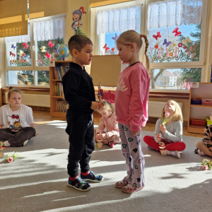 Dziewczynka składa chłopcu życzenia z okazji Dnia Mężczyzny i wręcza upominek - drewnianą rakietę z lizakiem