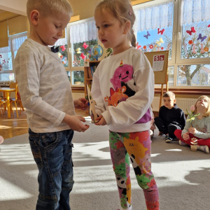 Dziewczynka składa chłopcu życzenia z okazji Dnia Mężczyzny i wręcza upominek - drewnianą rakietę z lizakiem