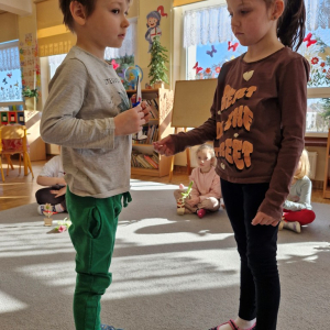 Dziewczynka składa chłopcu życzenia z okazji Dnia Mężczyzny i wręcza upominek - drewnianą rakietę z lizakiem