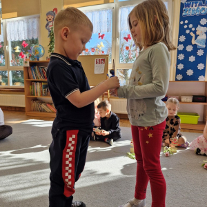 Dziewczynka składa chłopcu życzenia z okazji Dnia Mężczyzny i wręcza upominek - drewnianą rakietę z lizakiem