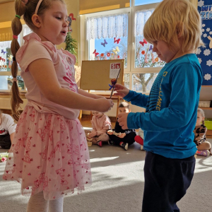 Dziewczynka składa chłopcu życzenia z okazji Dnia Mężczyzny i wręcza upominek - drewnianą rakietę z lizakiem