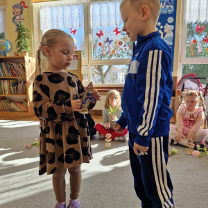 Dziewczynka składa chłopcu życzenia z okazji Dnia Mężczyzny i wręcza upominek - drewnianą rakietę z lizakiem