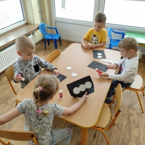 Dzieci siedzące przy stoliku przyklejają okrągłe płatki higieniczne na wcześniej wykonane tło na czarnej kartce tworząc bałwana