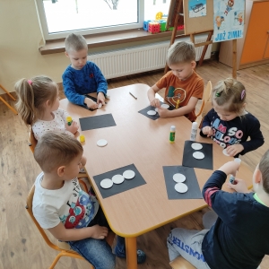 Dzieci siedzące przy stoliku przyklejają okrągłe płatki higieniczne na wcześniej wykonane tło na czarnej kartce tworząc bałwana