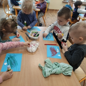 Dzieci siedzące przy stoliku kolorują szablon sanek i wyklejają tło białymi skrawkami papieru